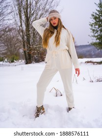 Winter Lifestyle, Young Blonde Caucasian Girl With White Winter Outfit And Woolen Hat In The Snow Next To Snowy Pine Trees, Holidays In Nature