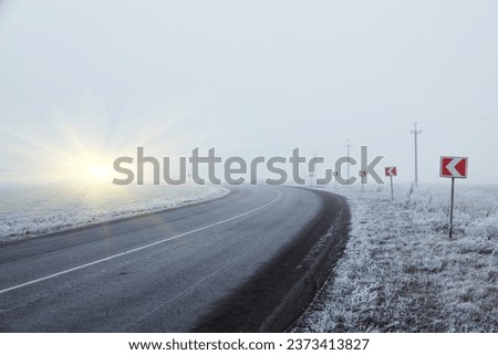 Winter morning on a winding road. The trees and ground are covered in a thick layer of frost. The road turning around a bend. A signpost is standing in front of the bend, warning of a sharp turn.