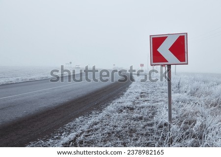 Winter morning on a winding road. The trees and ground are covered in a thick layer of frost. The road turning around a bend. A signpost is standing in front of the bend, warning of a sharp turn.