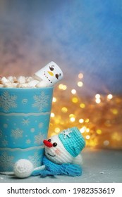 Winter Mug With Marshmellow Snowman Up And Knitted Handmade Stand Snowman In Blue Scarf Down Against Background Of Festive Lights