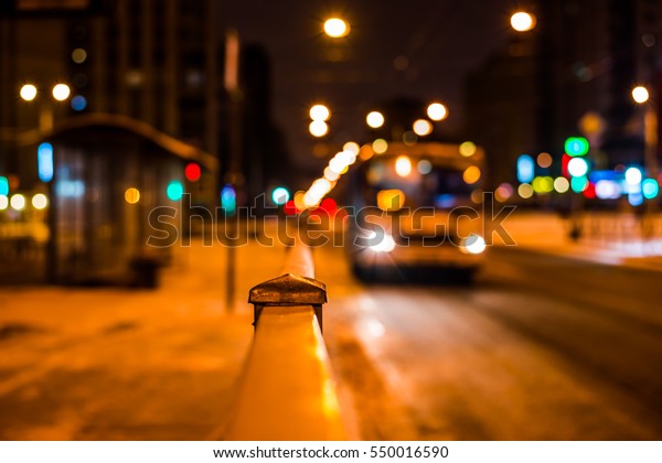 Winter night\
in the big city, the bus arrives at the bus stop. Close up view\
from the handrail on the sidewalk\
level