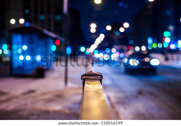 Winter night in the big city, the bus stop and the\
stream of cars. Close up view from the handrail on the sidewalk\
level, image in the blue\
tones