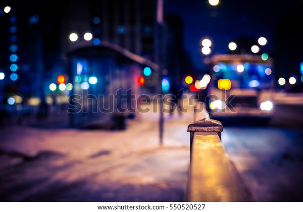 Winter night in the\
big city, the bus arrives to a stop for passengers. Close up view\
from the handrail on the sidewalk level, image vignetting and the\
yellow-blue toning