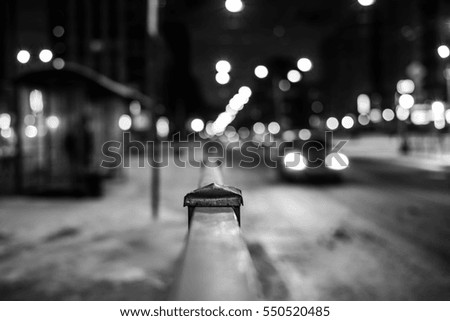 Winter night in the big city, the people waiting at bus stop next passing car. Close up view from the handrail on the sidewalk level, image in the black and white tones