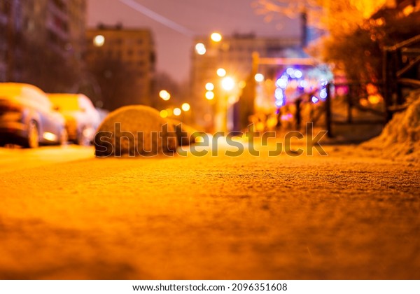 Winter in the night\
city. Sidewalk in the yard. Residential area. Snowy road. Parked\
cars. Working lights. Juicy colors. Focus on snow. Close up view\
from sidewalk level.