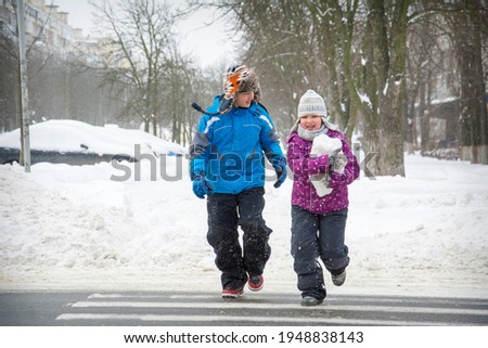 In winter, on the street in snowy weather, a brother and sister cross the road at a pedestrian crossing.