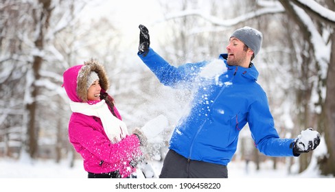 Winter outdoor fun happy interracial couple playing in snow throwing snowballs during snowball fight in park outside. Laughing asian woman with caucasian young man in winter coats.