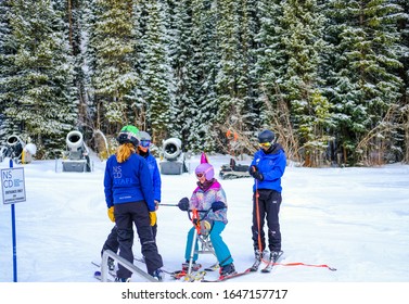 
Winter Park, Colorado/USA - 01/24/2020: Young Women Downhill Skiers Working For National Sport Center For Disabled Talking To  Disabled Girl On 3-ski Snow Bike; Snow Covered Trees In Background