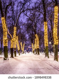 Winter Park At Night With Christmas Decorations, Lights, Pavement Covered With Snow And Trees.