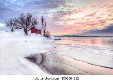 A winter photo of a red little cottage with some trees in the archipelago of Stockholm, Sweden. There is snow on the ground and some ice on the lake. The sky is colored from the beautful winter sunset