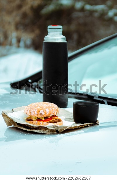 Winter picnic with tea\
and burger on the hood of a silver car against the background of\
the winter forest
