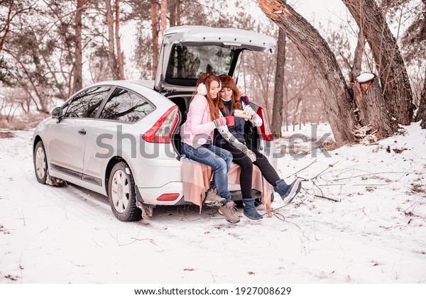 winter picnic together. Mom and student daughter\
are sitting in the car. Mother and daughter sit in the trunk of a\
car and drink tea from a thermos\
