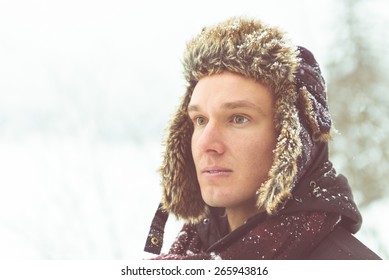 Winter Portrait Of An Attractive Young Man Outside In The Snow Wearing A Fur Bomber Hat.