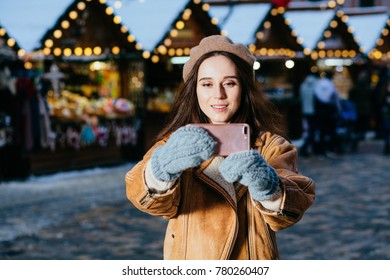 Winter Portrait Of Beautiful Hipster Woman Taking A Selfie Photo Outside On Winter, Snowy Day. Festive Christmas Fair On Background. Holidays, Christmas And People Concept.