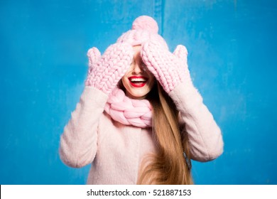 Winter portrait of a beautiful woman clothing her eyes with knitted gloves standing on the blue background