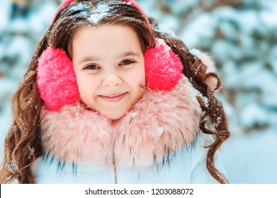 Winter Portrait Of Happy Kid Girl In Pink Earmuffs Walking Outdoor In Snowy Winter Forest. Happy Childhood And Active Winter Holidays Concept