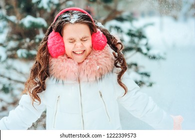 Winter Portrait Of Happy Kid Girl In Pink Earmuffs Walking Outdoor In Snowy Winter Forest. Happy Childhood And Active Winter Holidays Concept