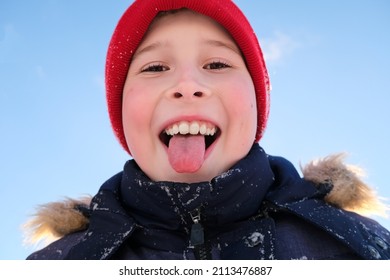 Winter portrait of a mischievous boy in close-up against the blue sky. A boy in winter clothes and a red knitted hat stuck out his tongue and catches snowflakes with his mouth.