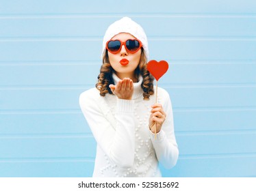 Winter portrait of woman with red heart shaped lollipop blowing her lips sending sweet air kiss wearing white hat, sweater on blue background - Powered by Shutterstock