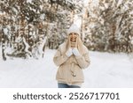 Winter portrait young beautiful woman with blond hair dressed in warm clothes with a knitted hat, smiling. Blurred snowy forest in the background