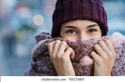 Winter portrait of young beautiful woman covering face with woolen scarf. Closeup of happy girl feeling cold outdoor in the city. Young woman holding scarf and looking at camera. - Shutterstock ID 705175807