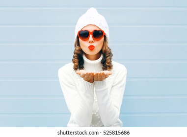 Winter portrait of young woman blowing her lips sending air kiss wearing white knitted sweater and hat, heart shaped sunglasses - Powered by Shutterstock