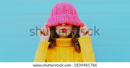 Winter portrait of young woman blowing red lips sending sweet air kiss female model wearing yellow knitted sweater on blue background