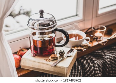 Winter Relax At Cozy Home. Glass Teapot With Hot Tea, Book, Glasses, Candle And Blanket By The Window, Snow Outside. Peaceful Hygge Atmosphere, Moody Light, Slow Life Philosophy Concept.