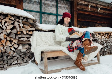 Winter Relax - Woman Sitting Outside A Cottage And Reading A Book On A Snowy Day. Woman Enjoying Time On Her Own On A Cold Winter Day.