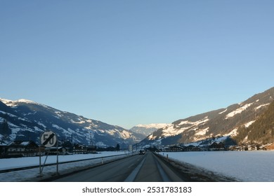 A winter road in perspective between small forested and snow-covered mountains under a clear blue sky. Cars drive in the distance