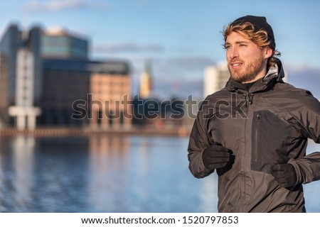 Winter running athlete man jogging outdoor in city outside wearing cold weather accessories - hat ,gloves , windproof sport jacket. Active healthy lifestyle.