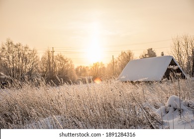 Winter Rural Landscape. Sunset In The Field. Tall Grass In A Snowdrift On The Background Of A Winter Sunset And An Abandoned Wooden House