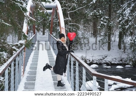 Winter scene, a beautiful girl with an inflatable red heart-shaped balloon on a bridge over a river in the Taevaskoja nature reserve.