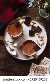 Winter Scene Flatlay Of Two Mugs Of Hot Chocolate On A Plate With Cardamom Pods And Holly, Woolen Gloves And A Scarf On A Dark Wood Background