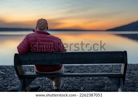Winter scene of a man with red jacket on a bench looking out over a lake at sunset.  Taking time for personal reflection, introspection, thinking about the past or the future.    Imagine de stoc © 
