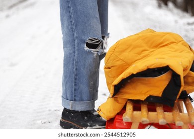 Winter scene with person in ripped jeans and black boots standing near a wooden sled with a yellow jacket on top. Snowy forest path on a cold winter day. Copy space - Powered by Shutterstock