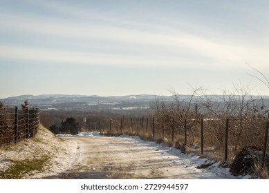 Winter scene of a snow-dusted path bordered by a fence. The distant hills and soft sunlight create a tranquil and serene atmosphere, ideal for nature walks or outdoor exploration.