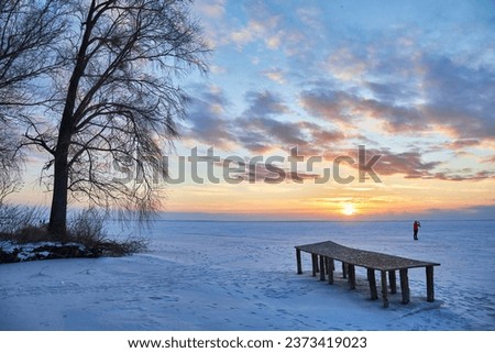 winter scene with a tranquil frozen lake. As the sun sets, the sky transforms into a masterpiece of vibrant colors, casting a warm, orange glow over the icy landscape and a charming wooden pier