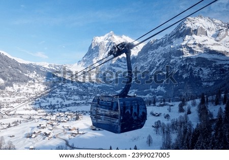 Winter scenery of the gondola of Eiger Express Cableway, which overlooks Grindelwald village on the snowy hillside and Wetterhorn mountain under blue sky in background, in Berner Oberland, Switzerland