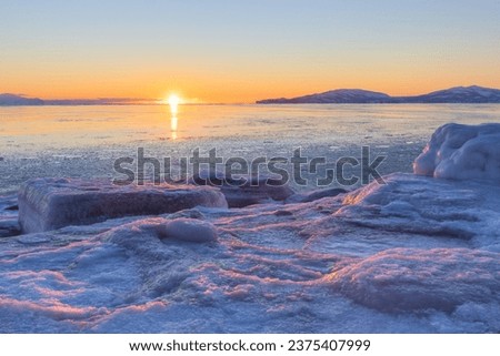 Winter seascape. Beautiful sunset. Ice-covered seashore and unfrozen water. Ice floes on the shore illuminated at sunset. Cold weather. Amazing northern nature. Coast of the Sea of Okhotsk, Russia.