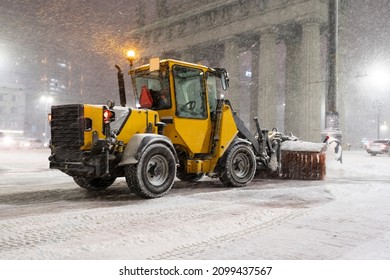 Winter Service Vehicle Clearing Thoroughfares Of Ice And Snow, Yellow Snow Plow Tractor Working On Empty Street At Night In Heavy Snowfall To Keep Roads Clear And Safe For Driving During Winter