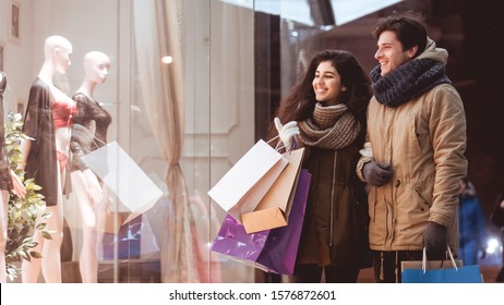 Winter Shopping. Happy Couple Standing Outdoor Of A Clothing Store And Pointing Finger At Shopwindow In Night City. Panorama