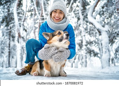 Winter Smiling Woman In Warm Hat And Scarf Hugging With Her Pet Welsh Corgi Dog, Having Fun In Snow At Winter Park