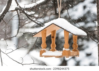 Winter. Snow is falling. A wooden bird feeder is hanging on a tree branch in the park. Feeding birds on harsh and cold days. Beautiful diffused light.
