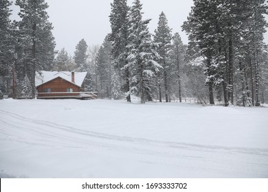 WInter Snowy Cabin House Exterior With Forest And Pine Trees And Snow Mobile Tracks.