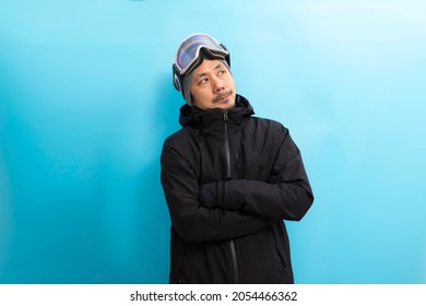 Winter Sport. Handsome Asian  Man With Snowboard Goggles And Black Anorak Coat With Curious Face Stand On Blue Background.