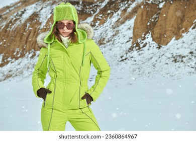 Winter sport in the mountains. Portrait of a fashionable girl posing in green ski suit against a snowy winter landscape. Winter fashion. 