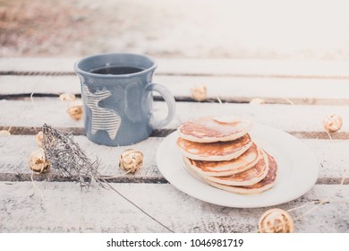 Winter still life on wooden table with hot blue mug of coffee, tea and pancakes with honey and butter surrounded with Christmas lights