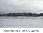 Winter in Stockholm reveals a tranquil view of frozen waters, with historic buildings atop a hill overlooking the icy expanse. Snow blankets the ground, creating a serene atmosphere.