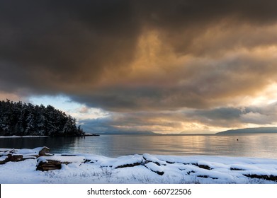 A Winter Storm In The San Juan Islands. Snow Covers The Coastline On Lummi Island In The San Juan Island Archipelago. Dramatic Clouds Cover The Sky All The Way To The Horizon.